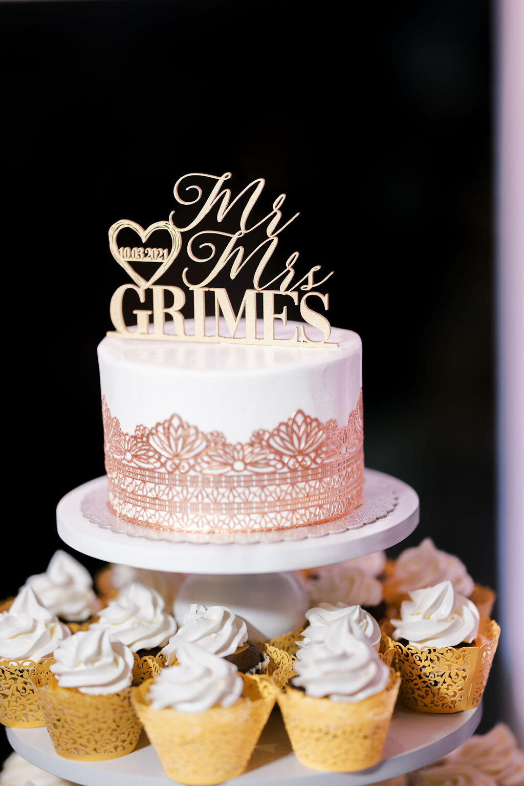 a white wedding cake decorated with an elegant pattern surrounded by cupcakes with gold liners and white frosting