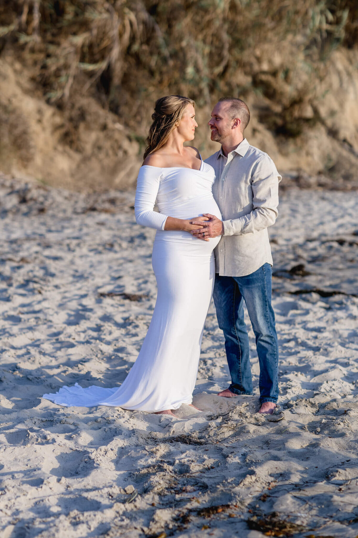 A pregnant woman in a white dress stands on a sandy beach, lovingly holding her belly. A man beside her, wearing a light shirt and jeans, gazes at her with a smile. The background features a rocky, plant-covered cliff.