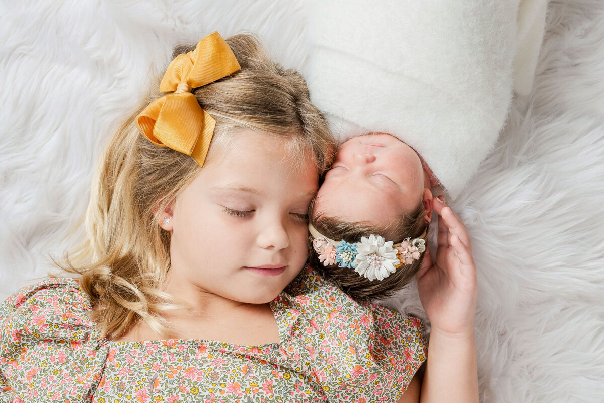 A big sister holding her newborn baby sister on her shoulder while laying on a white blankket.