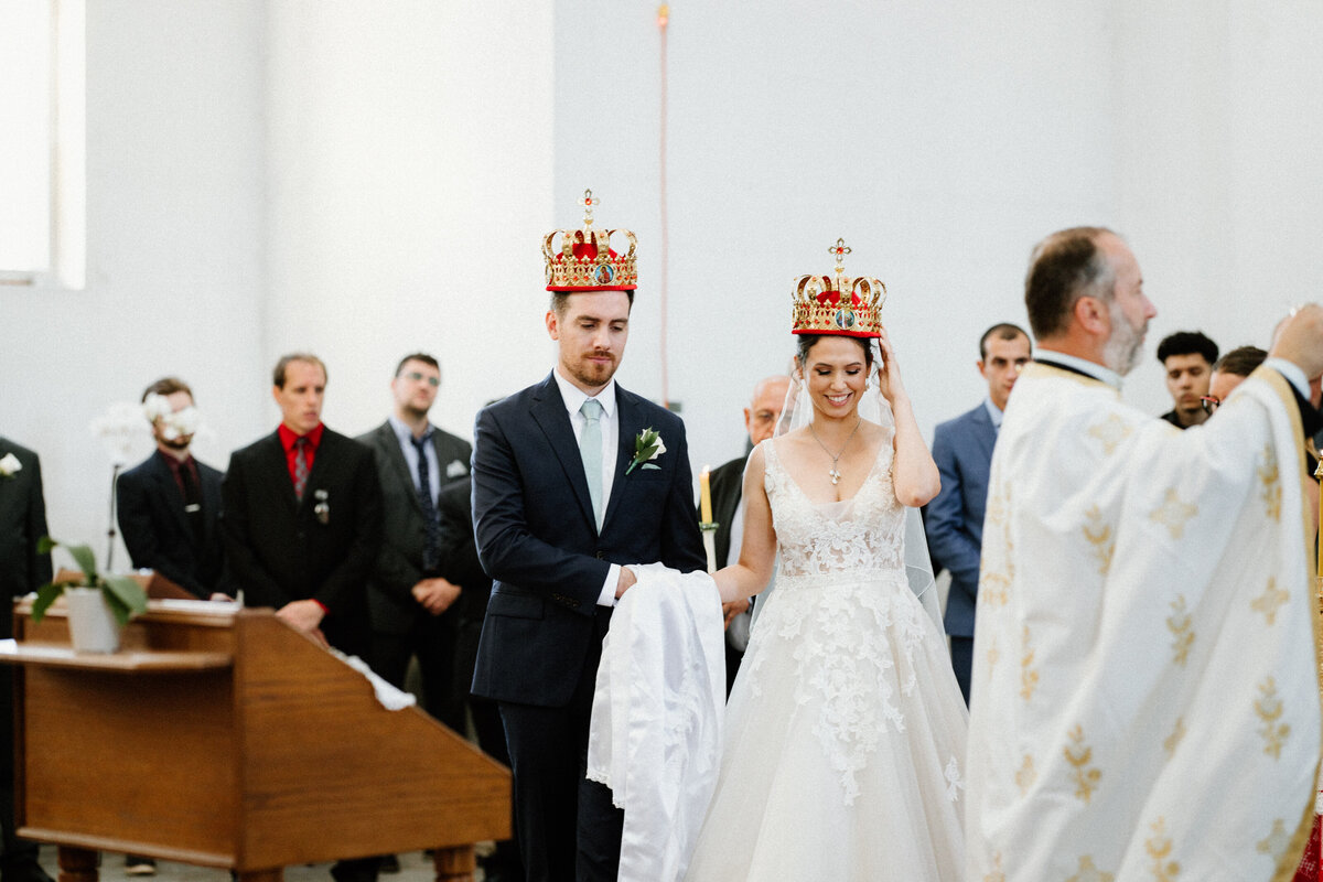Bride and groom hand in hand, while wearing a crown,as they listen to the pastor