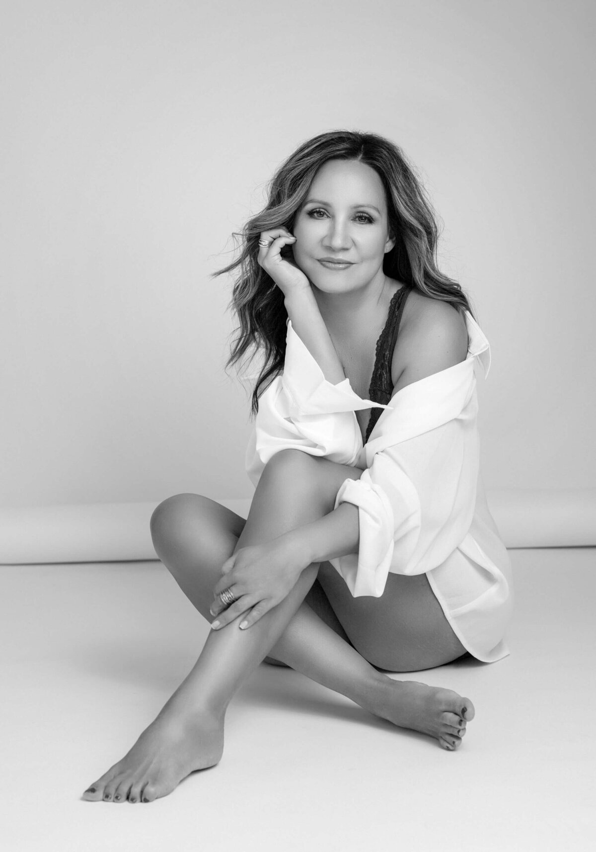Black and white portrait of a woman sitting on the floor, wearing an oversized white shirt, looking confidently at the camera.