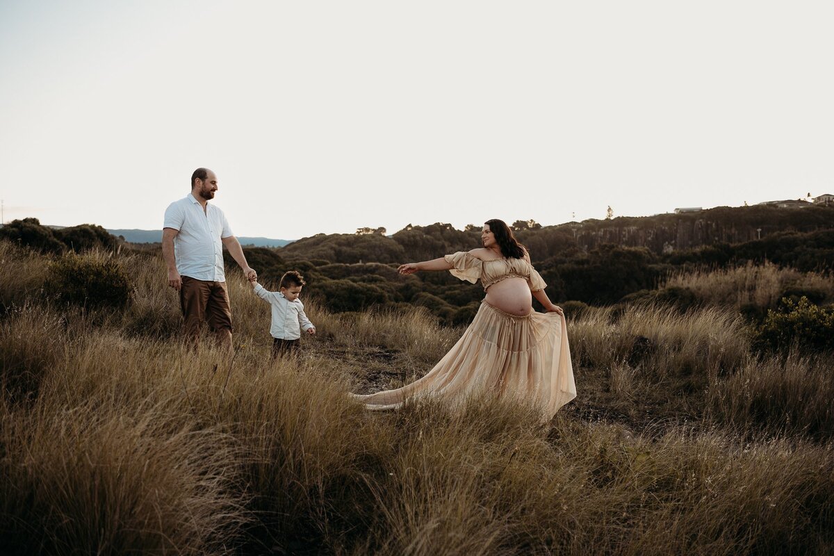 A pregnant woman in a flowing dress is extending her hand towards a man holding a child across a grassy field with hills in the background, perfectly capturing the essence of maternity photography showcased by an exceptional NSW family photographer.