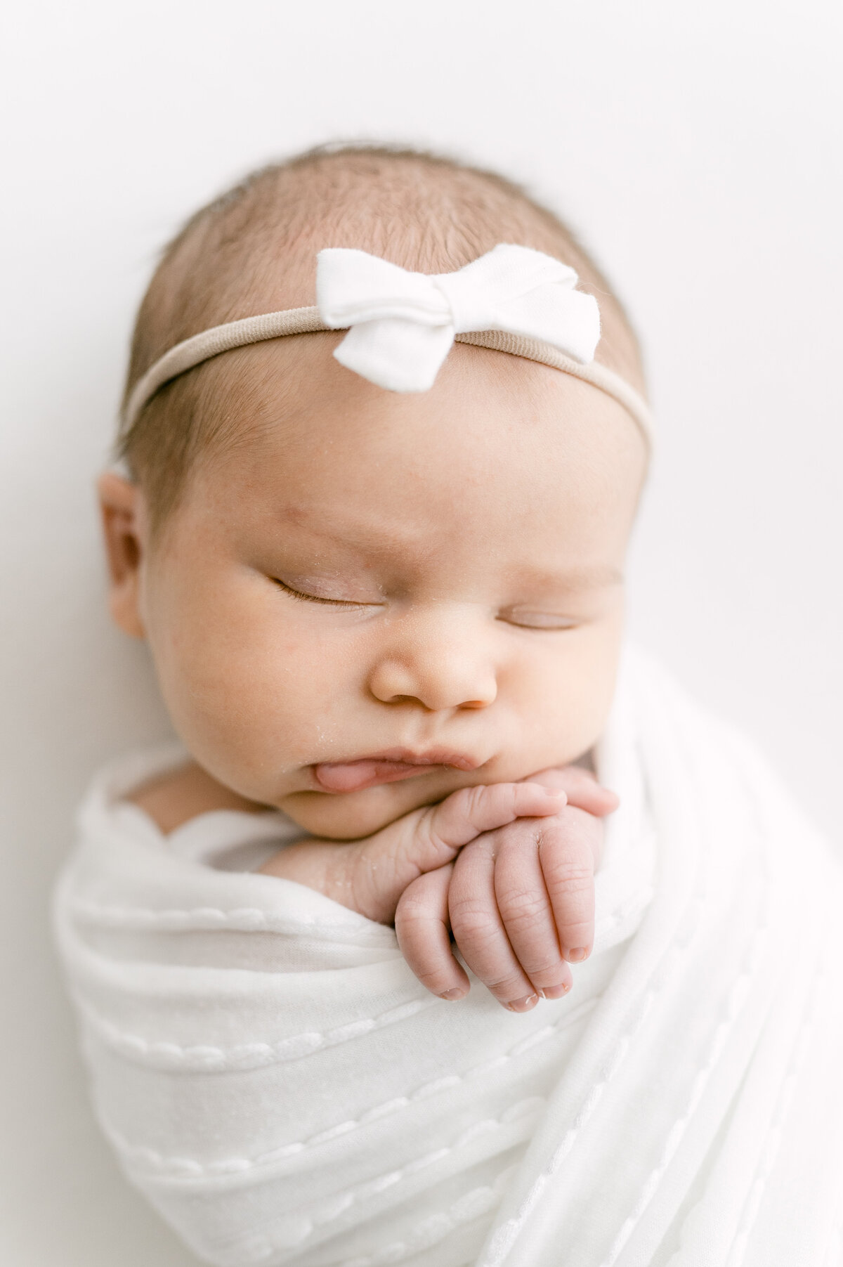 Close up photograph of a baby with a white bow and a white swaddle, hands out and under her chin.