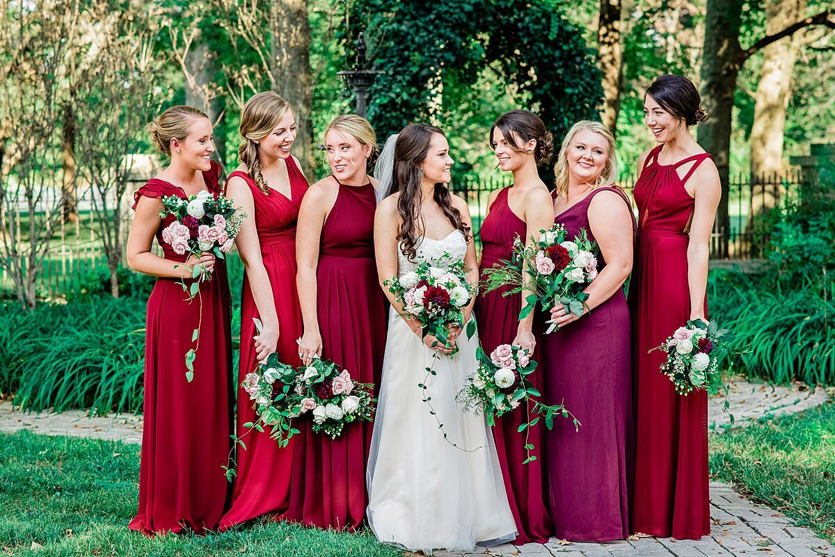 Bridesmaids wearing shades of red and standing in the courtyard at Historic Potter Barn holding large bouquets with ivy