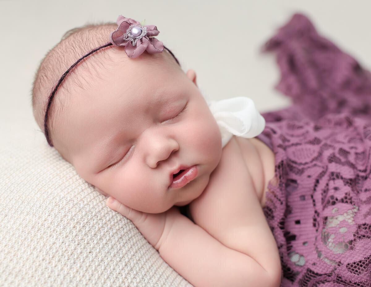 A peaceful newborn girl sleeping soundly on a tan backdrop, wrapped in a purple lace fabric. Captured in a newborn photography studio in Rochester, NY, showcasing the innocence and beauty of early life.