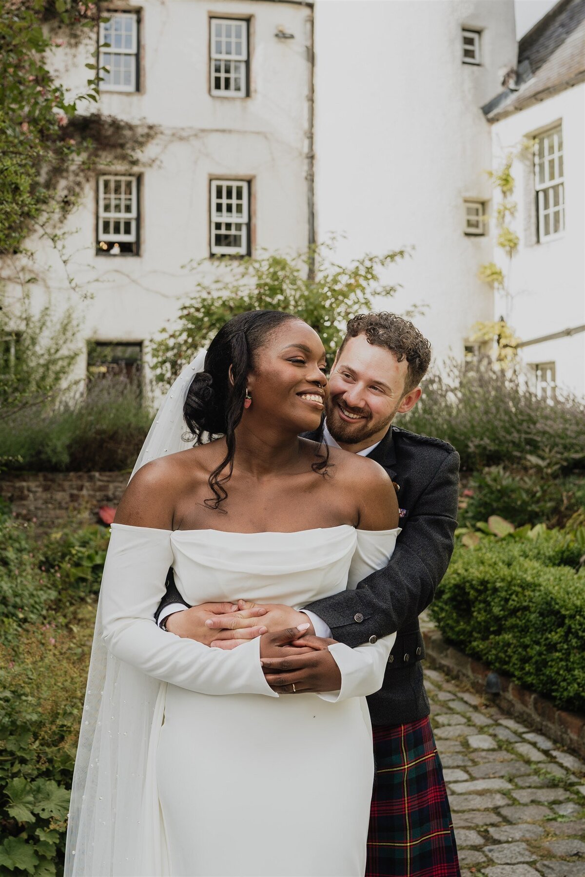 A groom wraps his arms around his wife from behind. They stand in a shaded courtyard and the white exterior of Logie Country House can be seen all around them. They stand on a cobbled surface and pose for wedding photos.