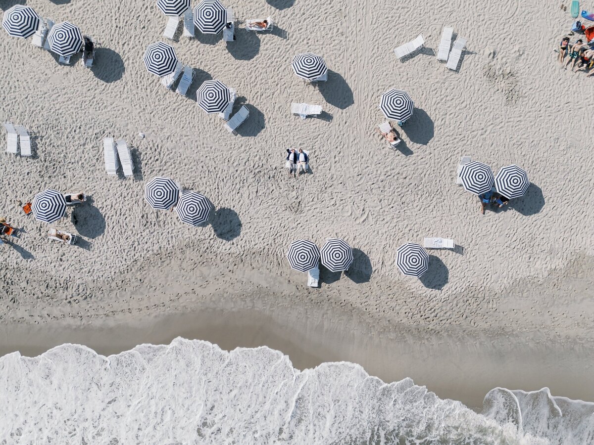 Drone portrait of teo grooms laying on the beach in cape may nj, surrounded by beach goers in matching rental umbrellas