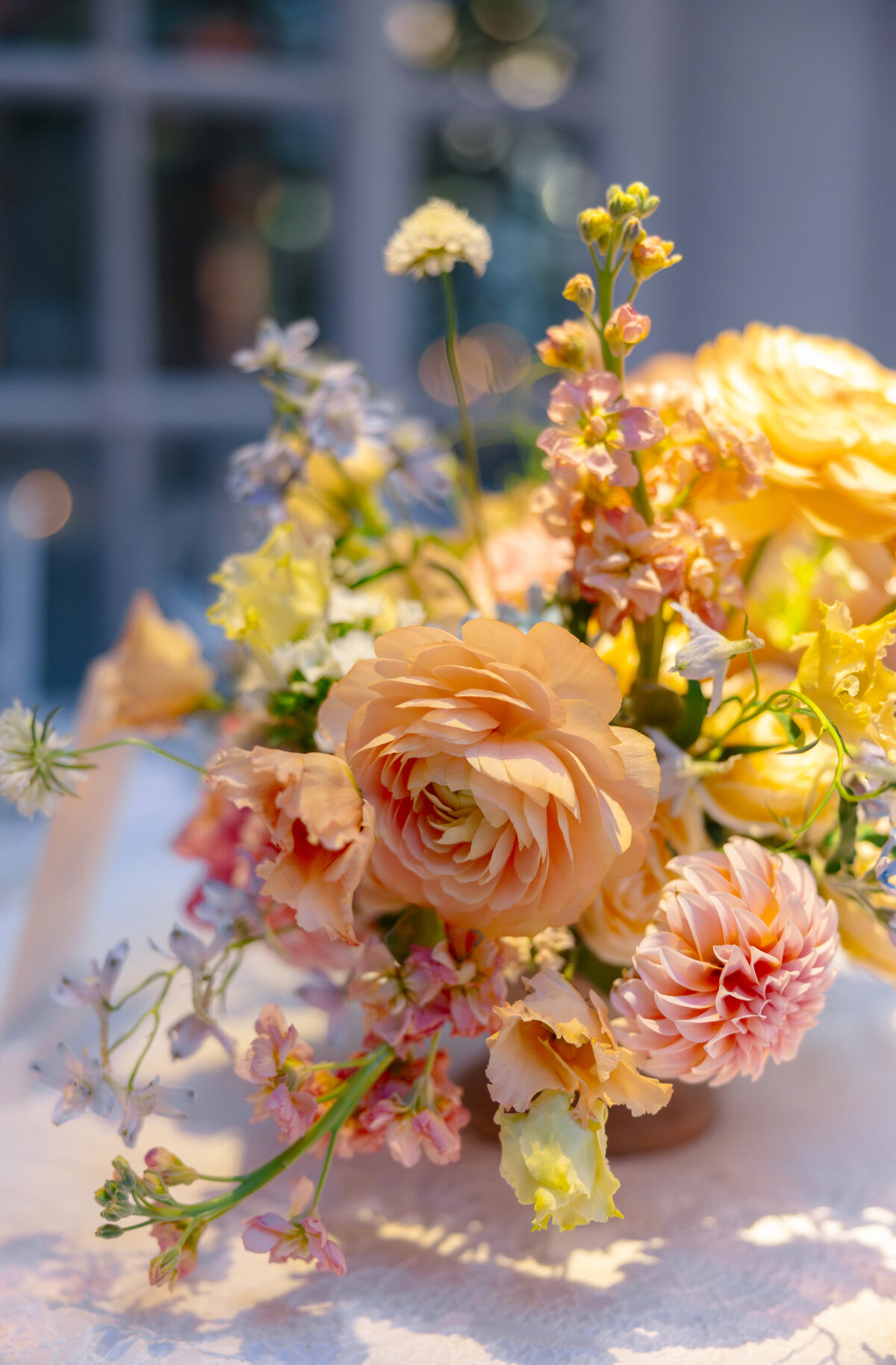 A vibrant floral arrangement featuring large peach and pink blooms, surrounded by smaller yellow and pastel flowers. The bouquet is set against a softly lit, blurred background, creating a warm and inviting atmosphere.