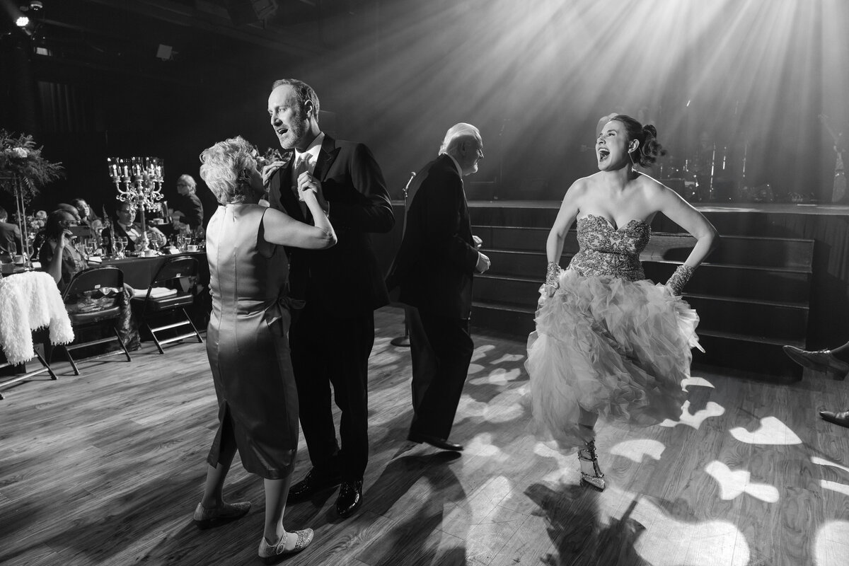Black and white photo of guests dancing under dramatic stage lighting at a theatrical wedding reception, with one guest in a sparkly, ruffled dress laughing and performing while others join in on the dance floor. Tables with candelabras and seated guests appear in the background, adding to the lively, cabaret-style ambiance.