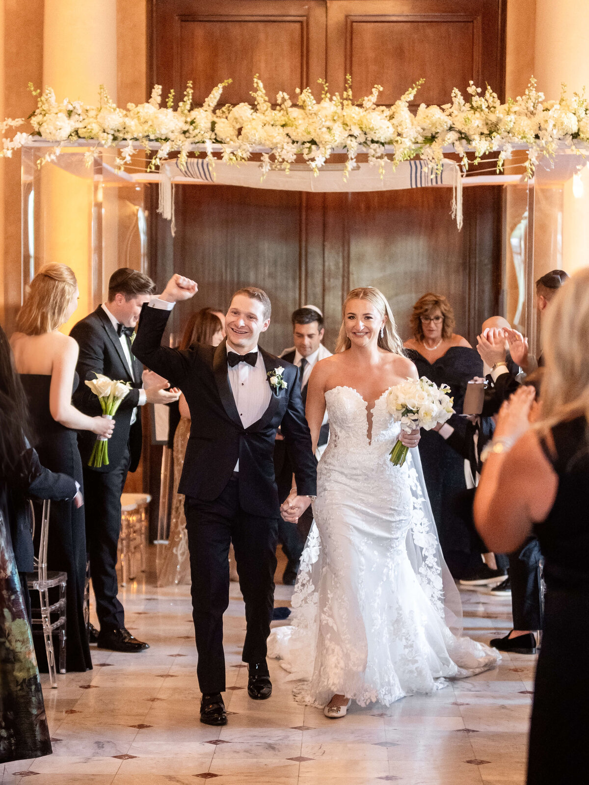 A newlywed couple walks down the aisle joyfully under a floral archway. The groom raises his fist in celebration, while the bride holds a bouquet of white flowers. Guests stand and clap in the elegant venue.
