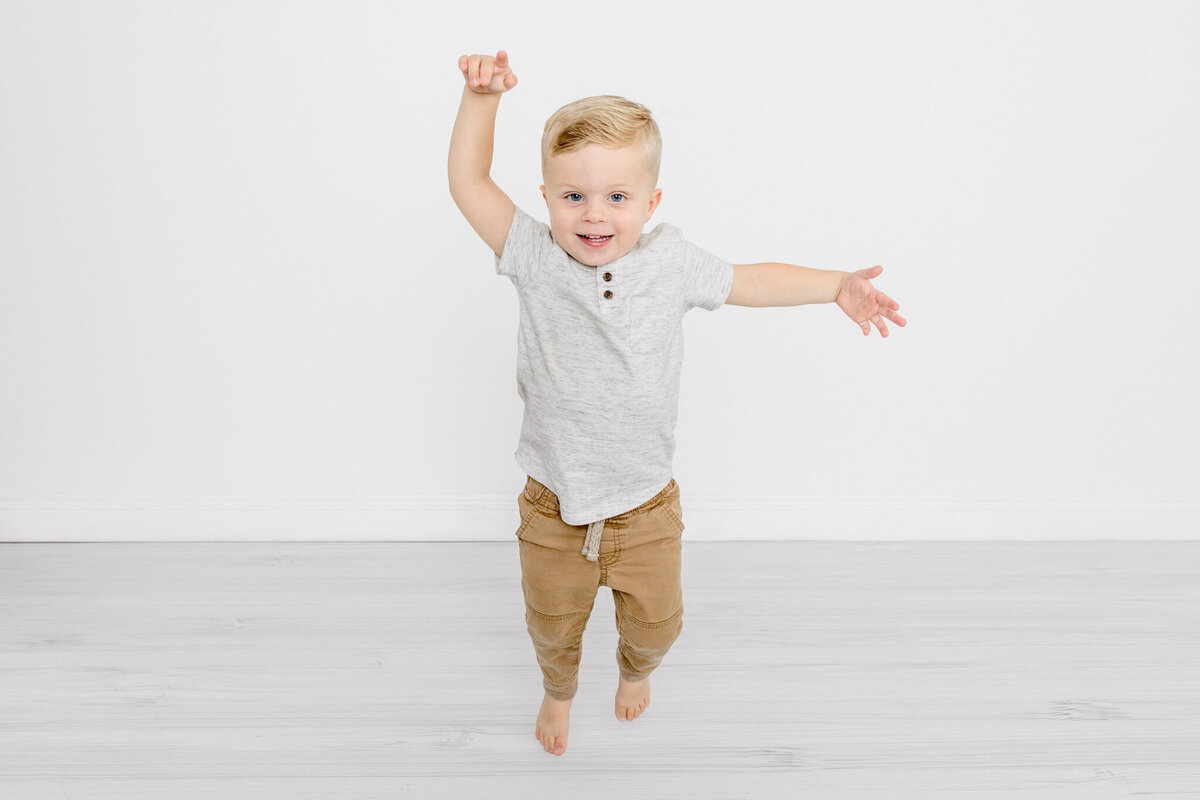 A little boy with blonde hair, wearing a grey shirt and brown pants, jumping in the air with his arms flailing and laughing against a white background.