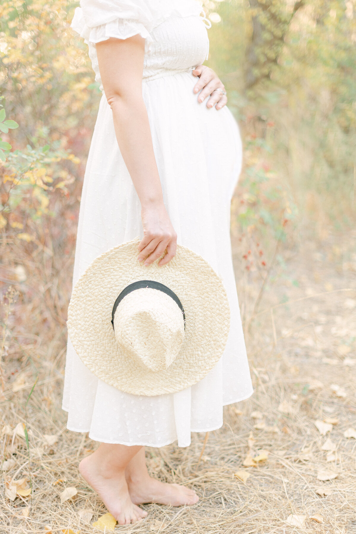 Portrait of a barefoot pregnant woman in a white summer dress with one hand on her belly and holding a hat out in the country