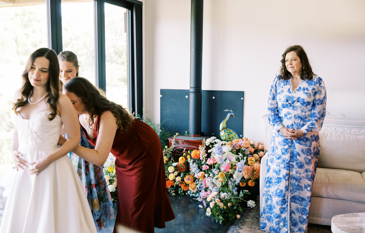 The mother of the bride looks on adoringly at her bride and the bridesmaids as they make final adjustments to her dress, standing in front of a beautiful backdrop of flowers, by film photographer My Sun and Stars Co.