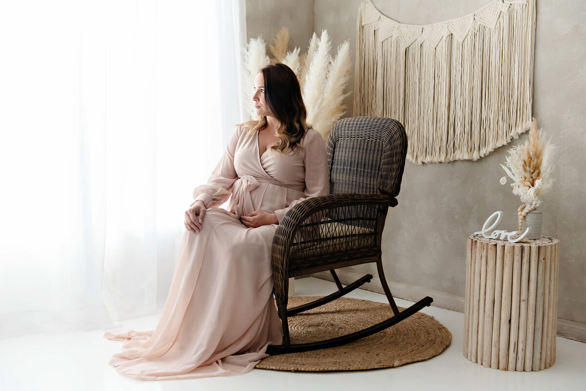 pregnant woman wearing a pink maternity dress sitting on a rocking chair for her studio maternity photography session in Burlington, Ontario