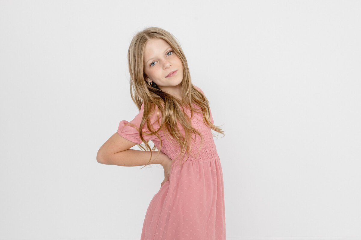 A pre-teen girl with long, blonde wavy hair wearing a light pink dress with her hands on her hips leaning back a bit and looking sassy at the camera in front of a white background.