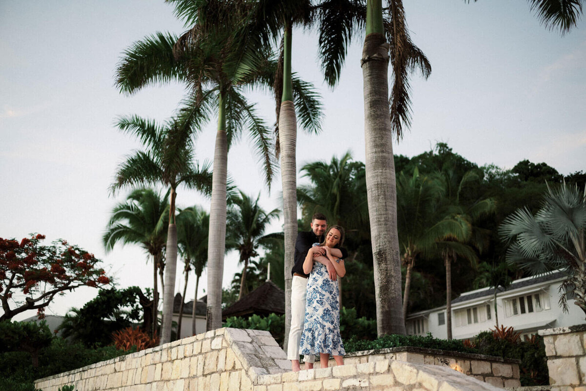 The engaged couple is standing on top of a low stone brick wall in front of Round Hill Hotel and Villas, Jamaica.
