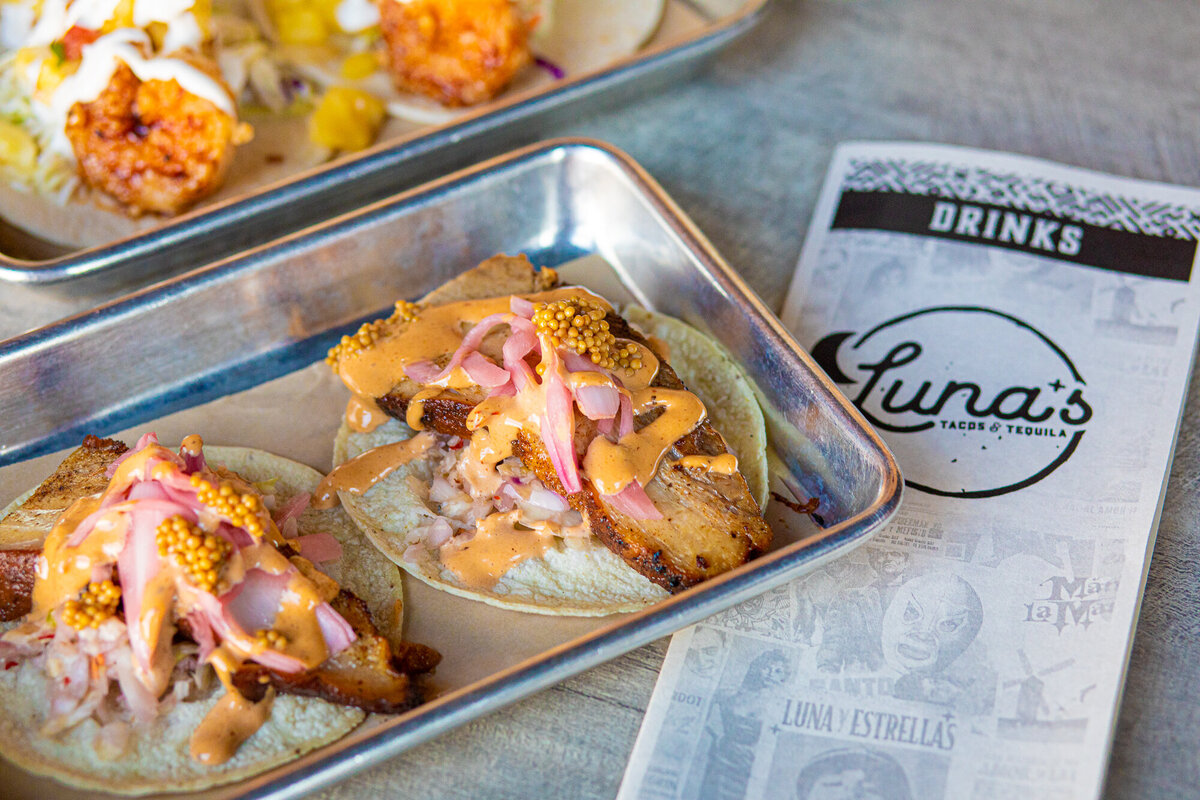 A plate of pork belly tacos is displayed next to a menu at Luna's Tacos and Tequila in Windsor, Colorado.