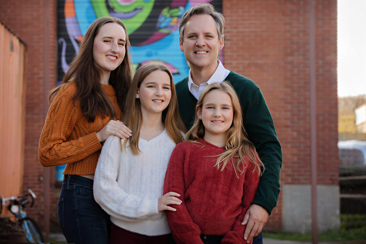 Proud dad embracing his three girls against a bright, multicolored mural at Camp North End Charlotte NC