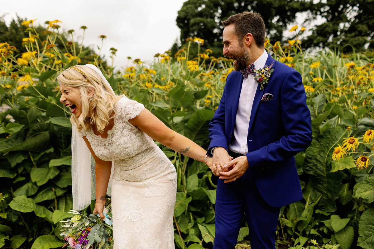 Bride and groom laughing candidly at each other stood in front of yellow flowers at Broughton Sanctuary