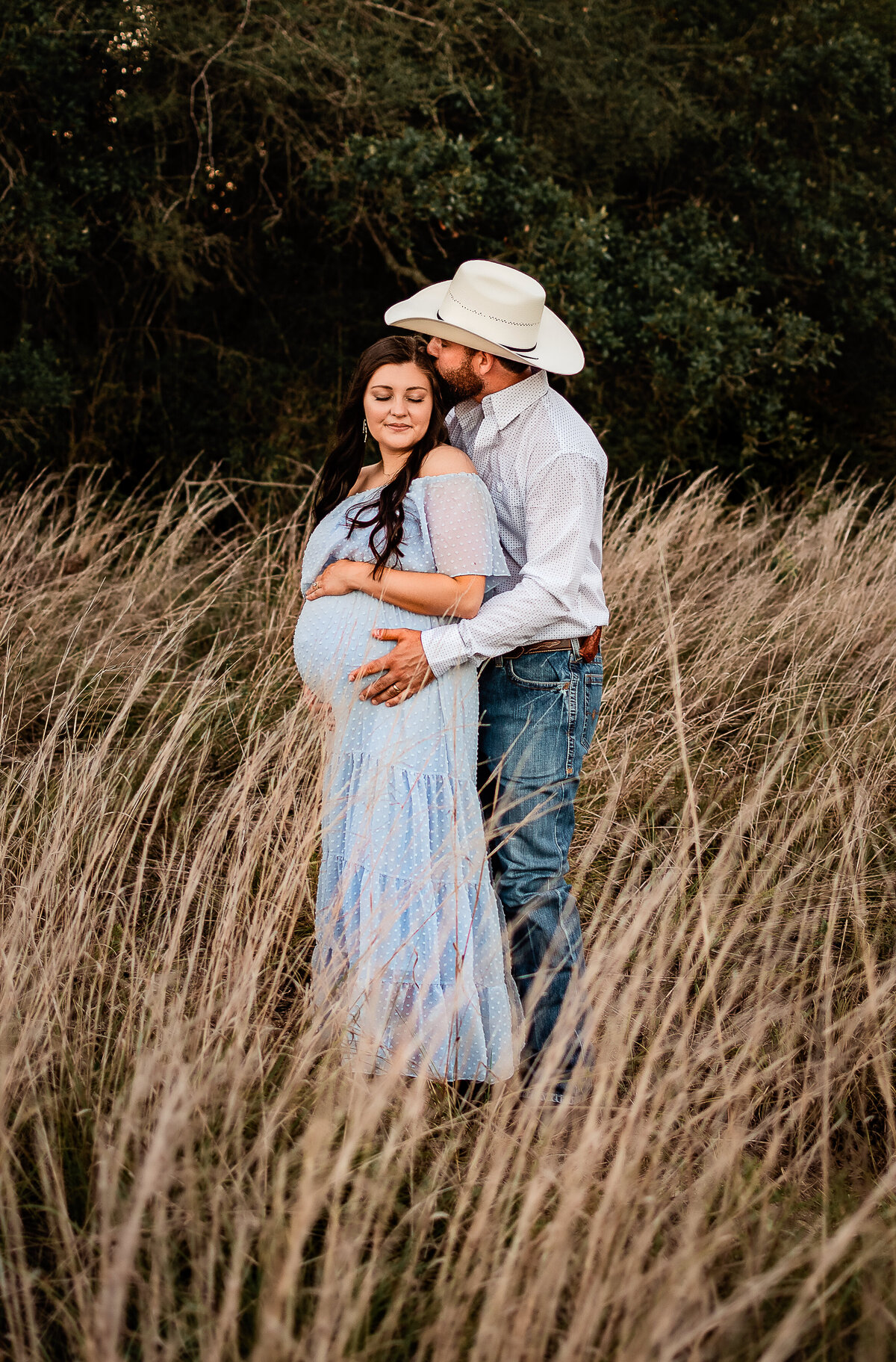 A dad to be hugs his pregnant wife from nehind and kisses her forehead in a field of long grass.