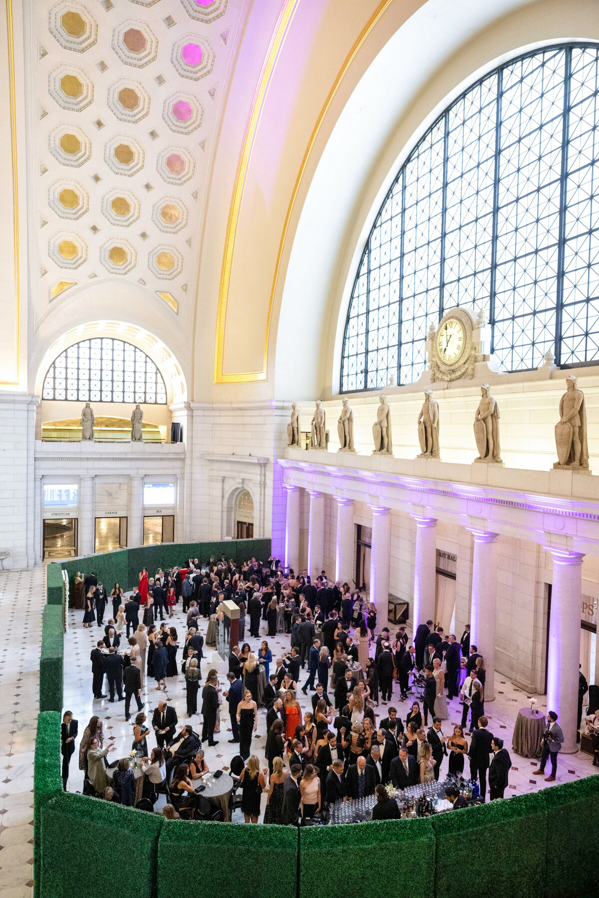 A large crowd gathers in the grand hall of a historic building with an arched ceiling and large windows. The space is adorned with statues and decorative patterns. People are mingling and socializing, surrounded by green hedges.