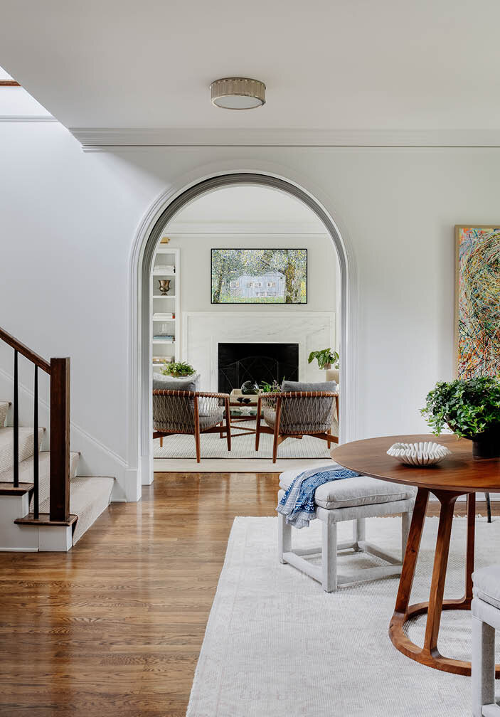 Discover a spacious foyer in a historic Hingham farmhouse, featuring a mid-century modern walnut table, benches, neutral rug, and custom art against gallery-like white walls.