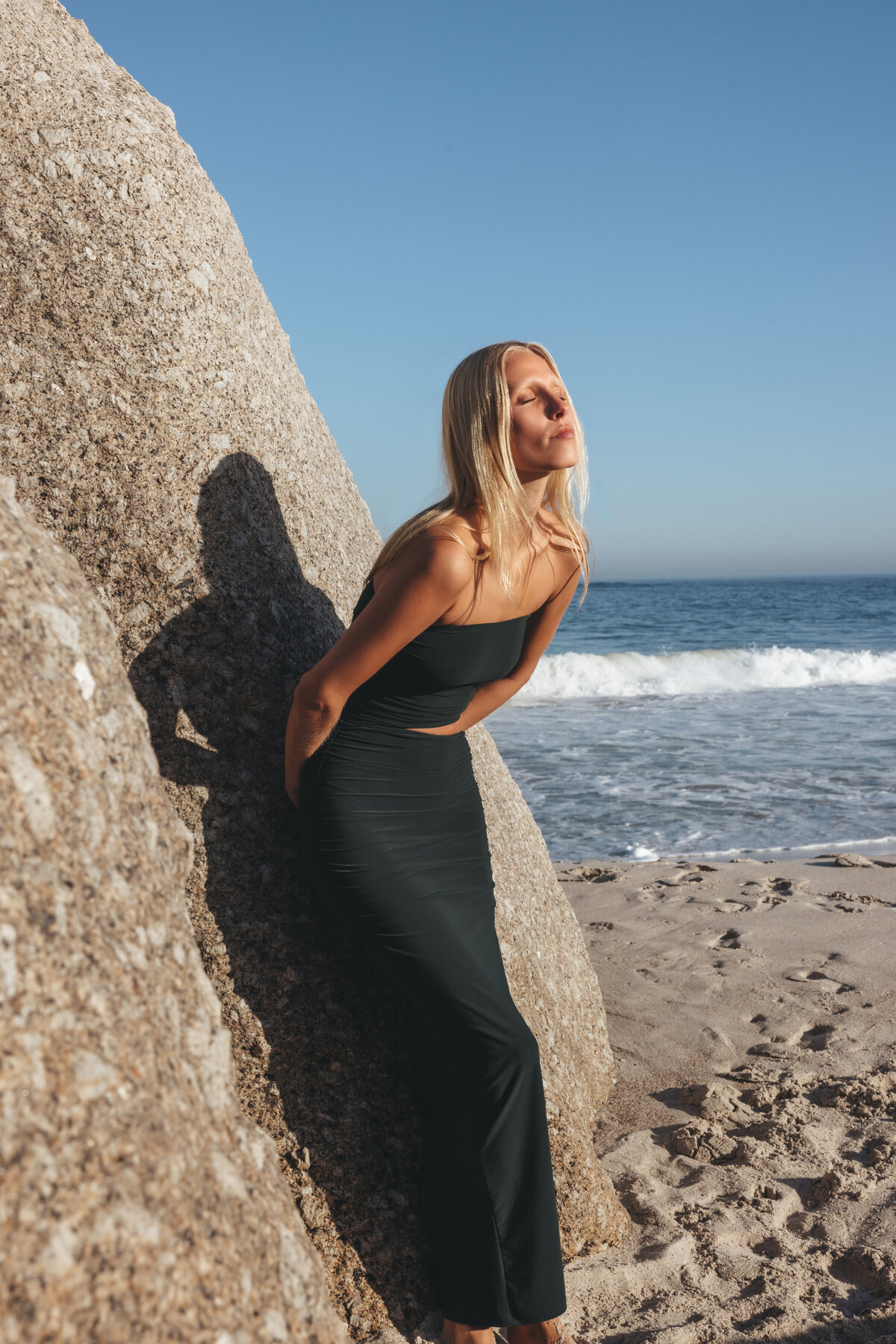 Model Johanna leunt tegen een grote rots aan het strand in Kaapstad, gehuld in een elegante zwarte strapless jurk, met haar gezicht gericht naar de zon en op de achtergrond de branding van de oceaan.