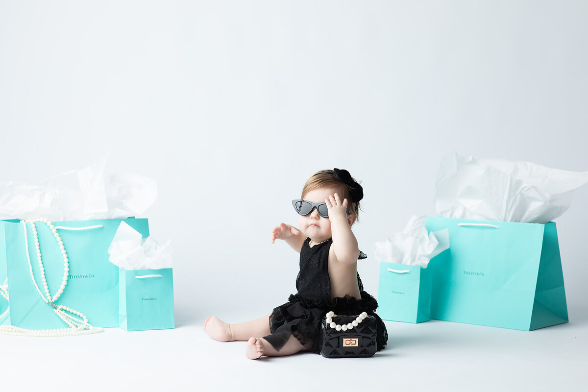 Dallas milestone photographer captures a baby girl in sunglasses and a black lace outfit, striking a pose with Tiffany blue gift bags and a toy purse adorned with pearls.