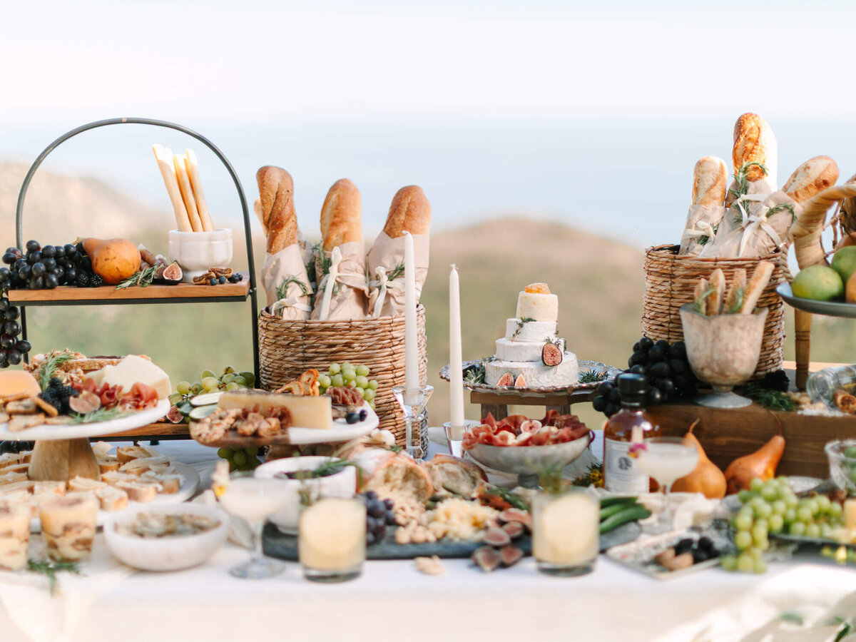 A beautifully arranged picnic table set outdoors, featuring artisanal bread, cheeses, fruits, and candles.
