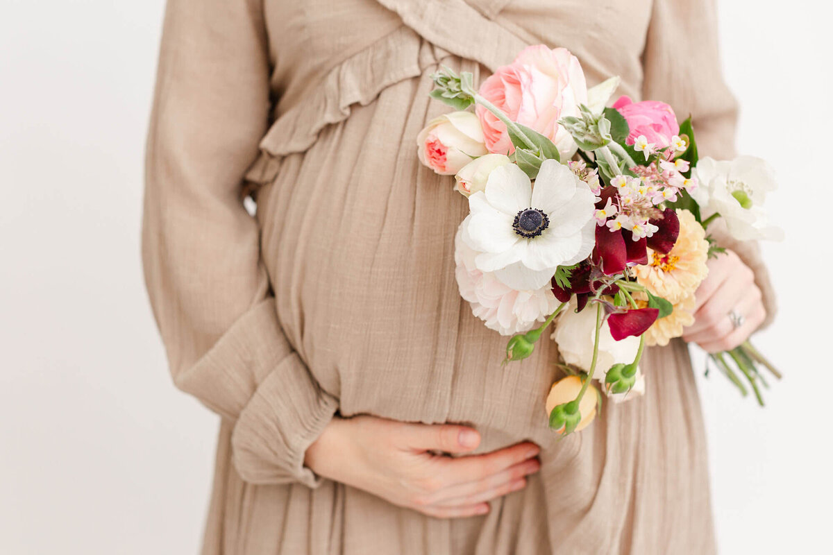 Close up image of pregnant belly. Mom is holding belly with one hand and a bouquet of flowers in the other hand. Studio maternity photo session in Portland, Oregon.