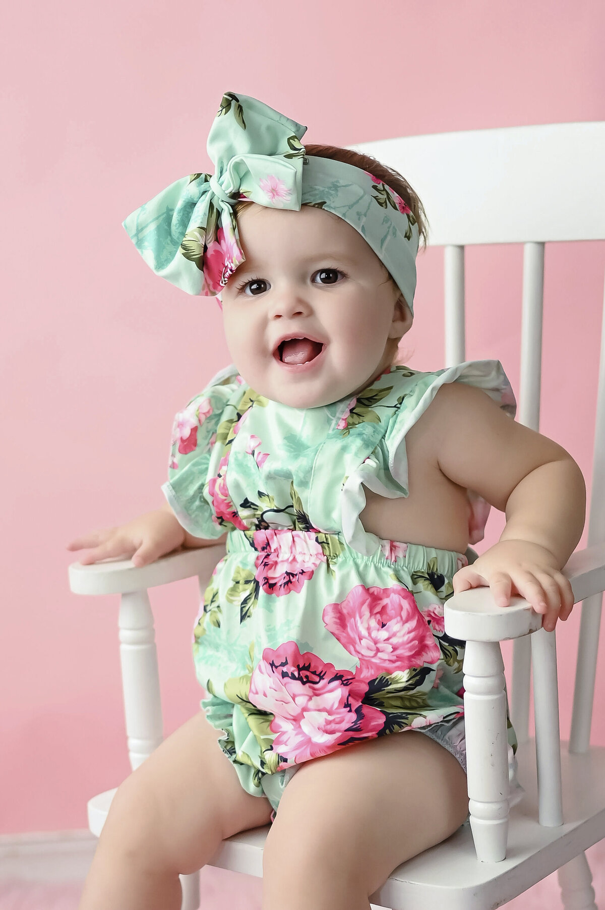 Toddler in floral dress and matching headbow sitting on a white chair against a pink background.