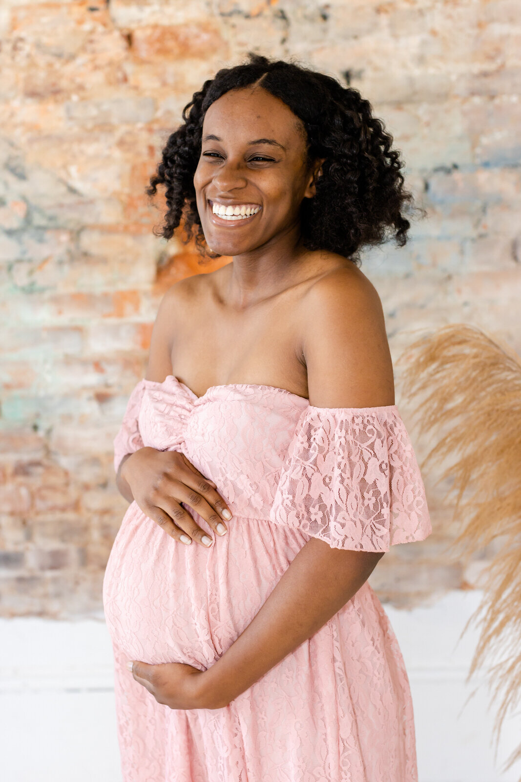 Mom smiles and holds onto baby bump in front of brick wall at Relic15 Studio in Buford