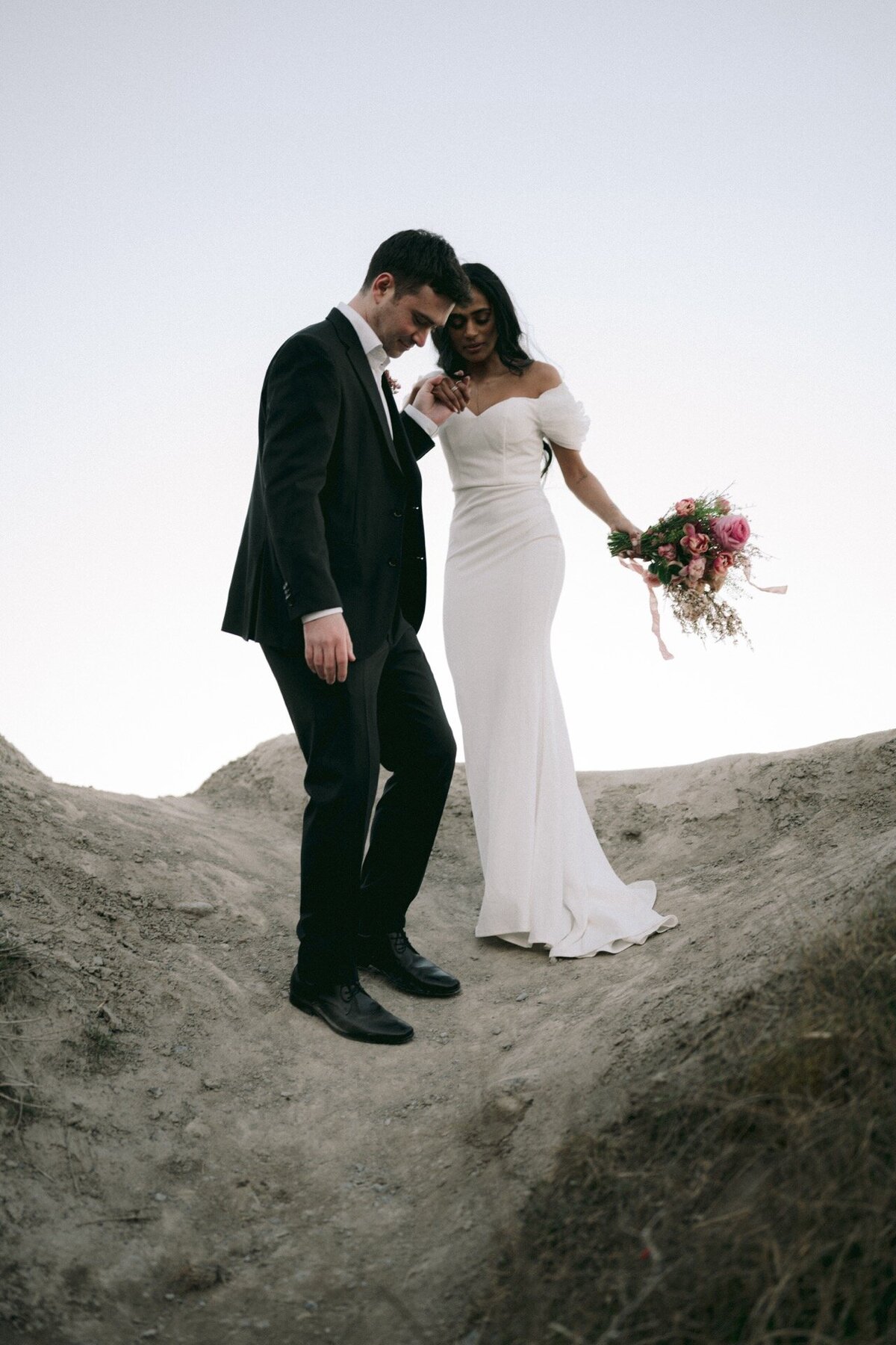 Groom helps his bride in high heel from climbing to the top of Sunrise Hill.