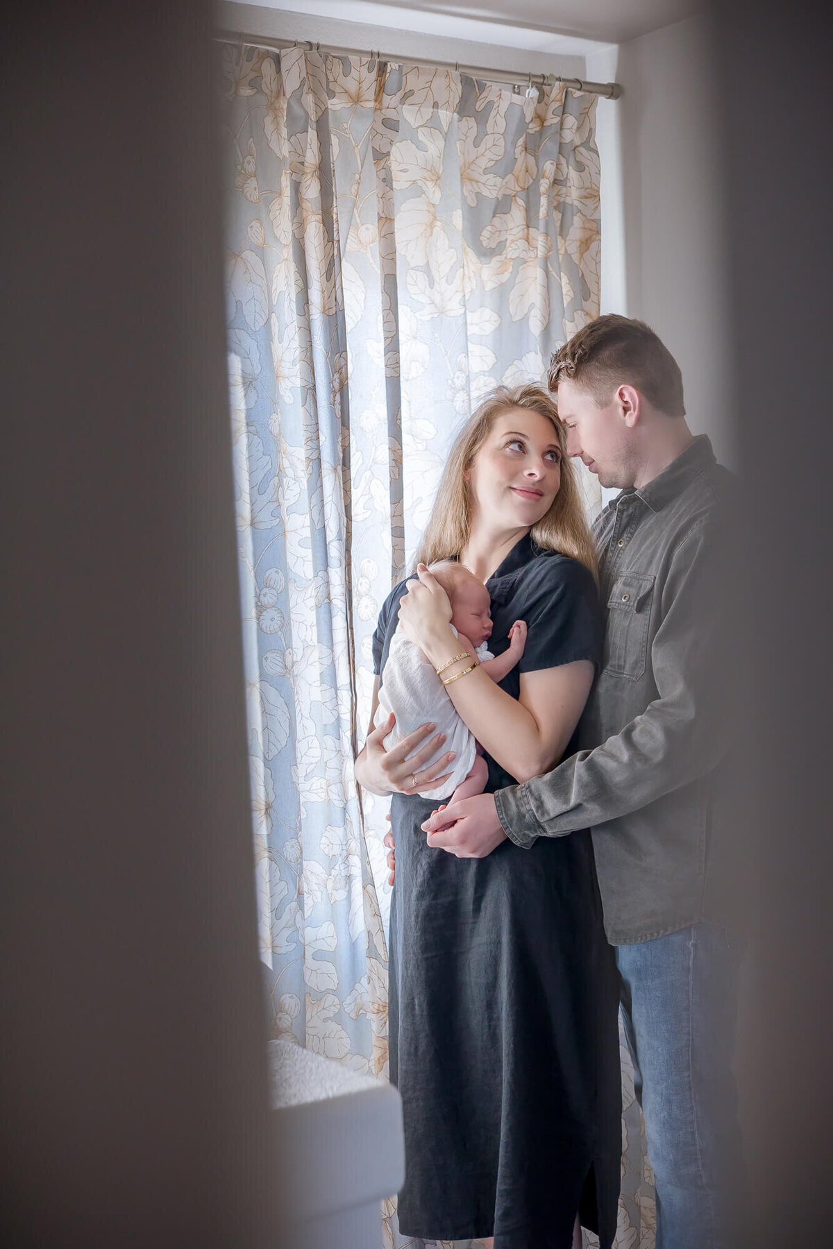 Couple standing by a window looking at each other with their newborn son during their photo session with Jessica Bowles