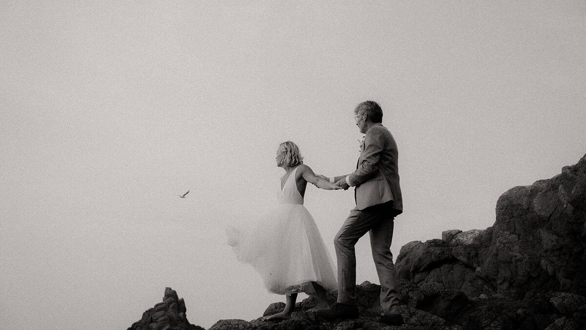 Patrick accompagne Gwladys sur les rochers en lui tenant le bras. Cliché en noir et blanc signé Kamescop, vidéaste et photographe mariage vintage.