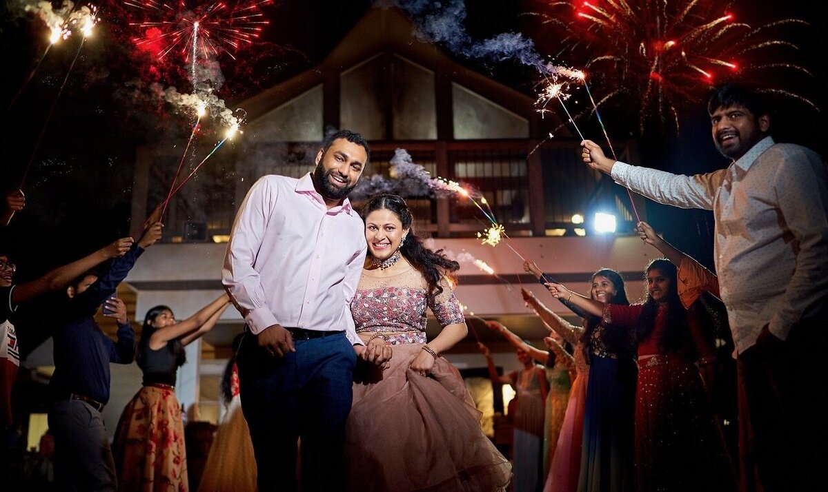 Amidst a vibrant celebration, a couple smiles and holds hands as friends surround them with sparklers. They stand in front of a Leesburg VA wedding venue, fireworks adding flair to the night—a perfect setting for those seeking charming wedding packages in Virginia.