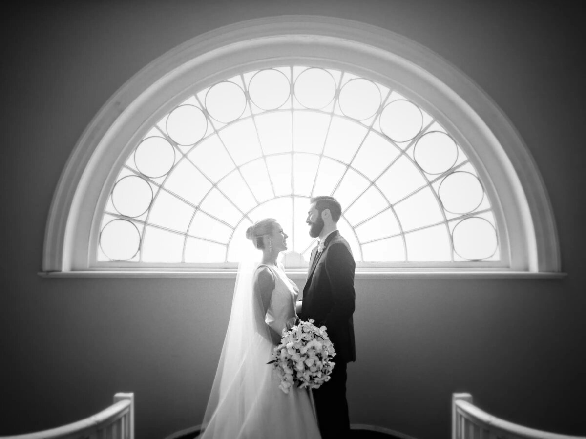 A bride and groom stand beneath an elegant arched window, their silhouettes framed by the natural light.