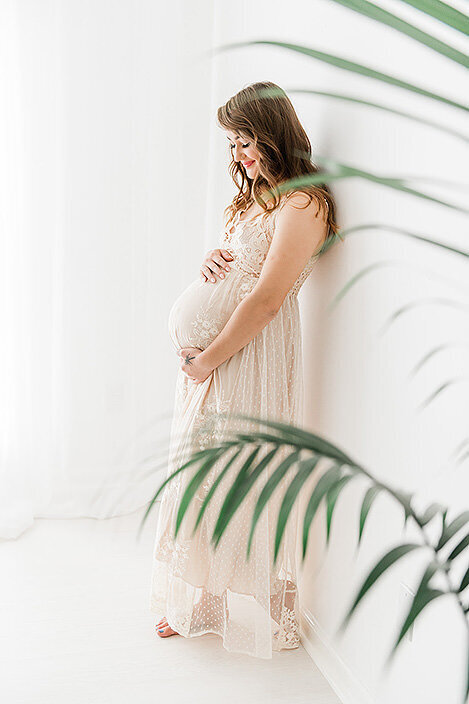Smiling expecting mother in a neutral gown, cradling her bump while standing by a window with palm leaves in the foreground.