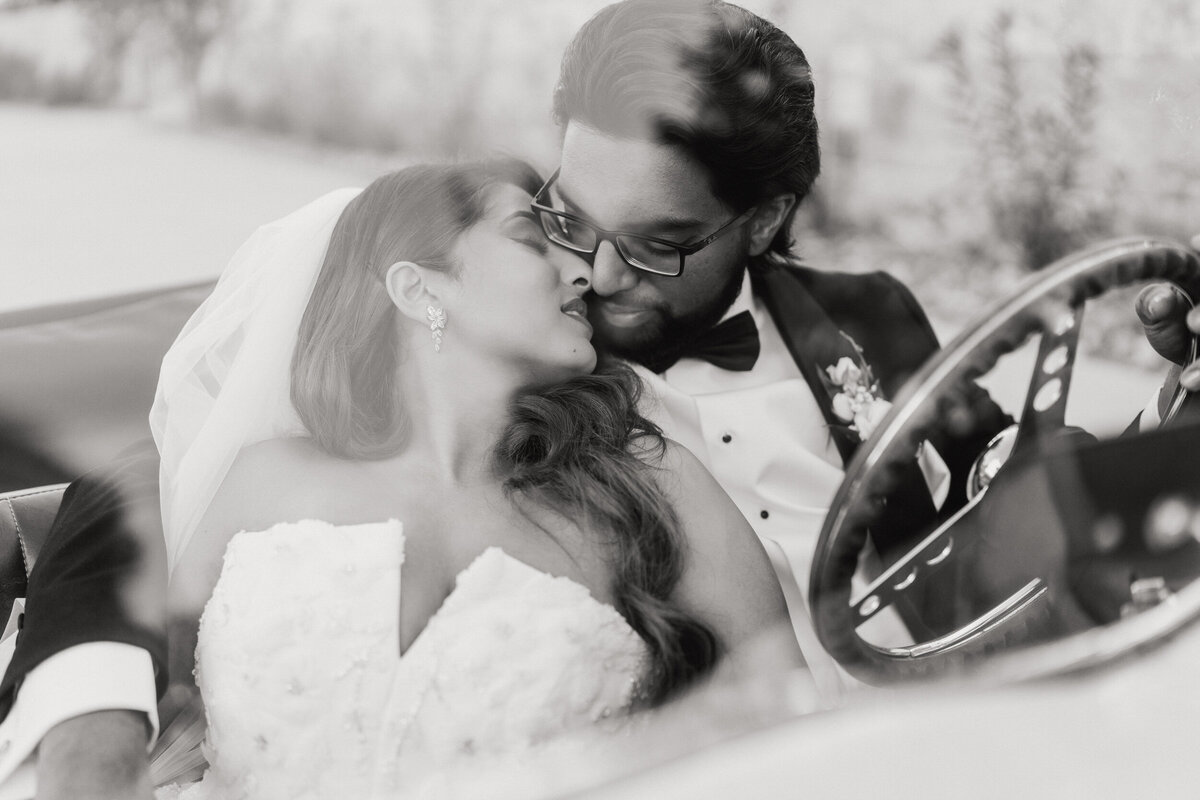 A black and white photograph of Heera and Nithin on their wedding day at The Preserve at Canyon Lake in Canyon Lake, Texas. Bride and groom are sitting in a vintage Rolls Royce, he’s in the driver’s seat and she’s leaning back against him. Her head is turned towards him as they move in close to kiss. Wedding photography by Stacie McChesney of Vitae Weddings.