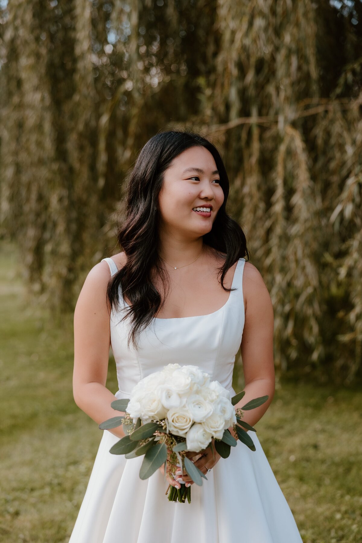 A bride stands in a lush garden at Stagecoach Inn, holding a bouquet of white roses and eucalyptus leaves. She wears a simple, elegant gown with a square neckline, her dark hair styled in loose waves. The soft natural light and the greenery in the background create a serene and joyful moment as she smiles off-camera.