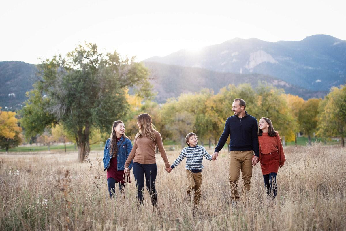 A family of 5 walk while laughing and holding hands through a field of tall golden grass at sunset