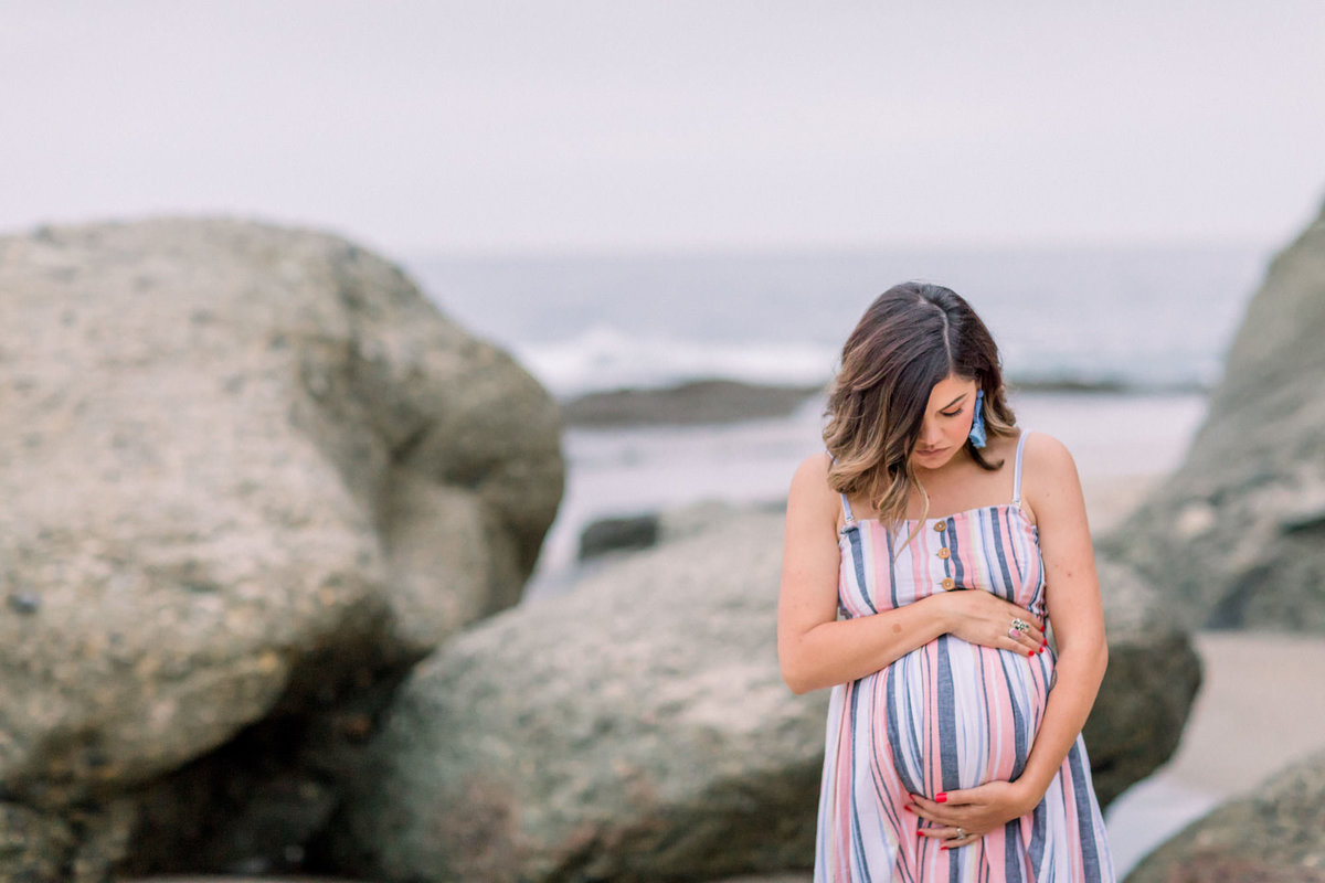 Pregnant woman holds her stomach while posing amongst the rocks on the beach