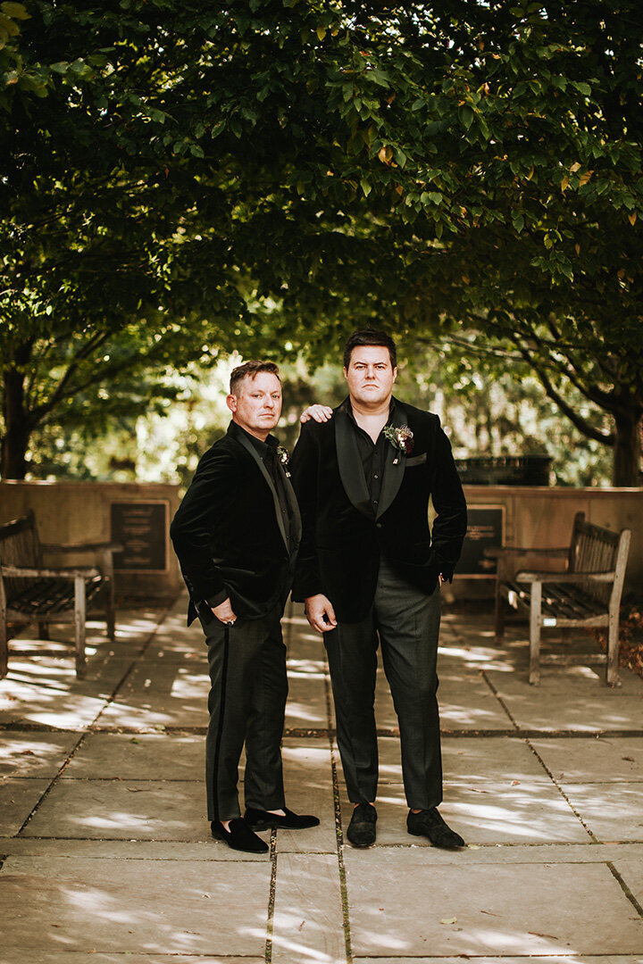 Two grooms wearing black tuxedos pose side by side in an outdoor quart yard.