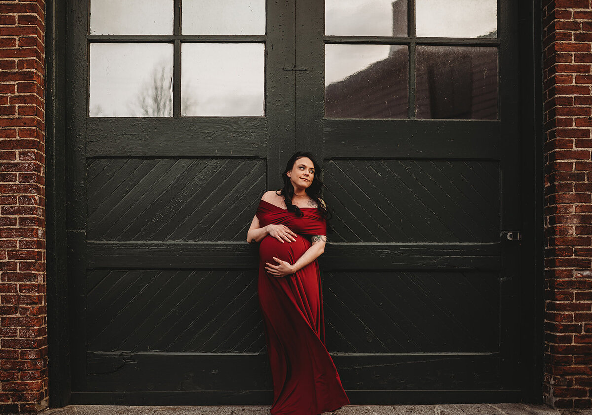 Family photographers Maryland photographs outdoor maternity session with pregnant woman in a dark red maternity dress holding her bump while standing in front of a wood garage on a red brick building