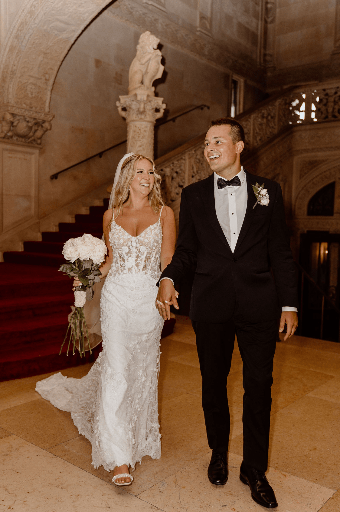Newlyweds joyfully walk hand in hand down an ornate staircase, the bride in a stunning lace gown holding a bouquet of white roses, and the groom in a classic black tuxedo. The grand architectural details and elegant setting provide a timeless backdrop for this candid, celebratory moment.
