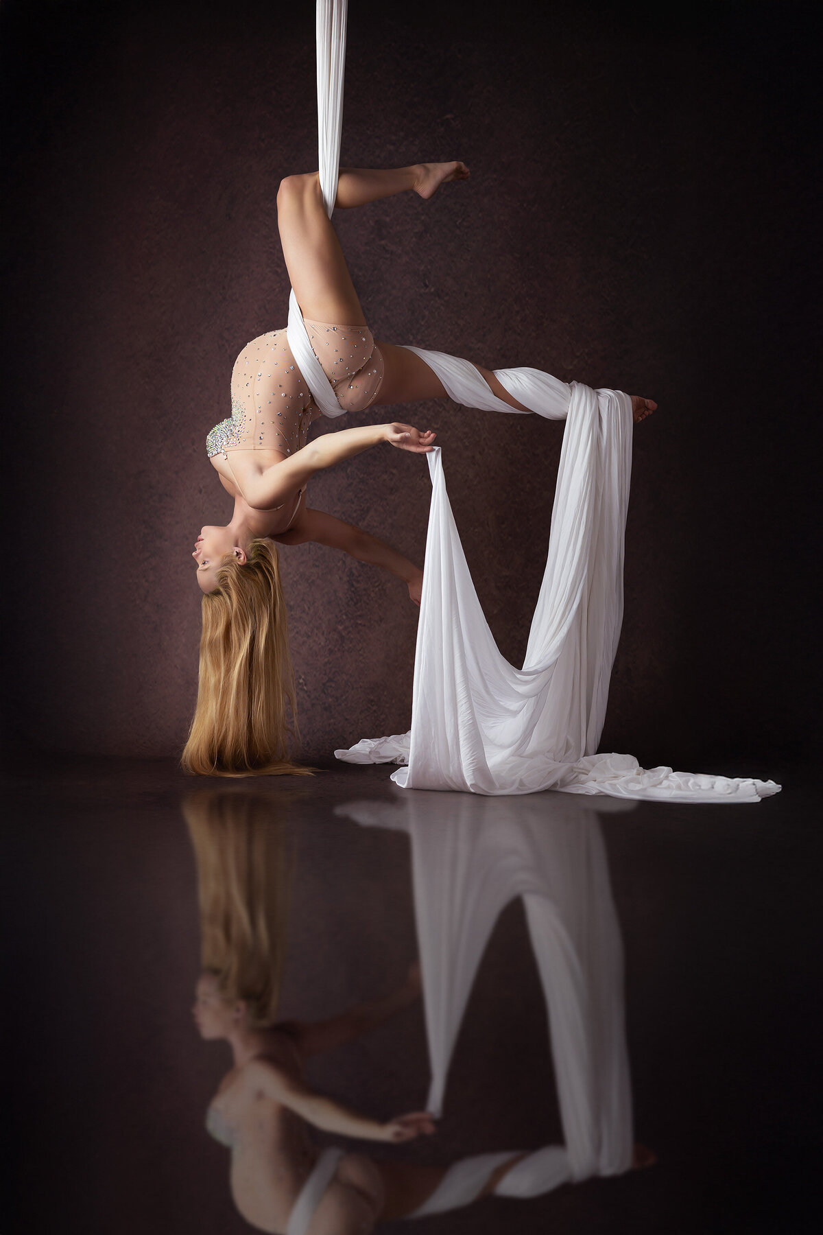 A woman elegantly suspended upside down from white silk, her long hair cascading toward the floor while her sparkling bodysuit shimmers against a deep, rich backdrop. The silks flow around her, creating a dynamic composition captured by a talented DFW maternity photographer.