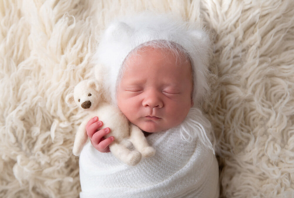 newborn baby boy wrapped in white wearing a white bear hat and holding a teddy bear.