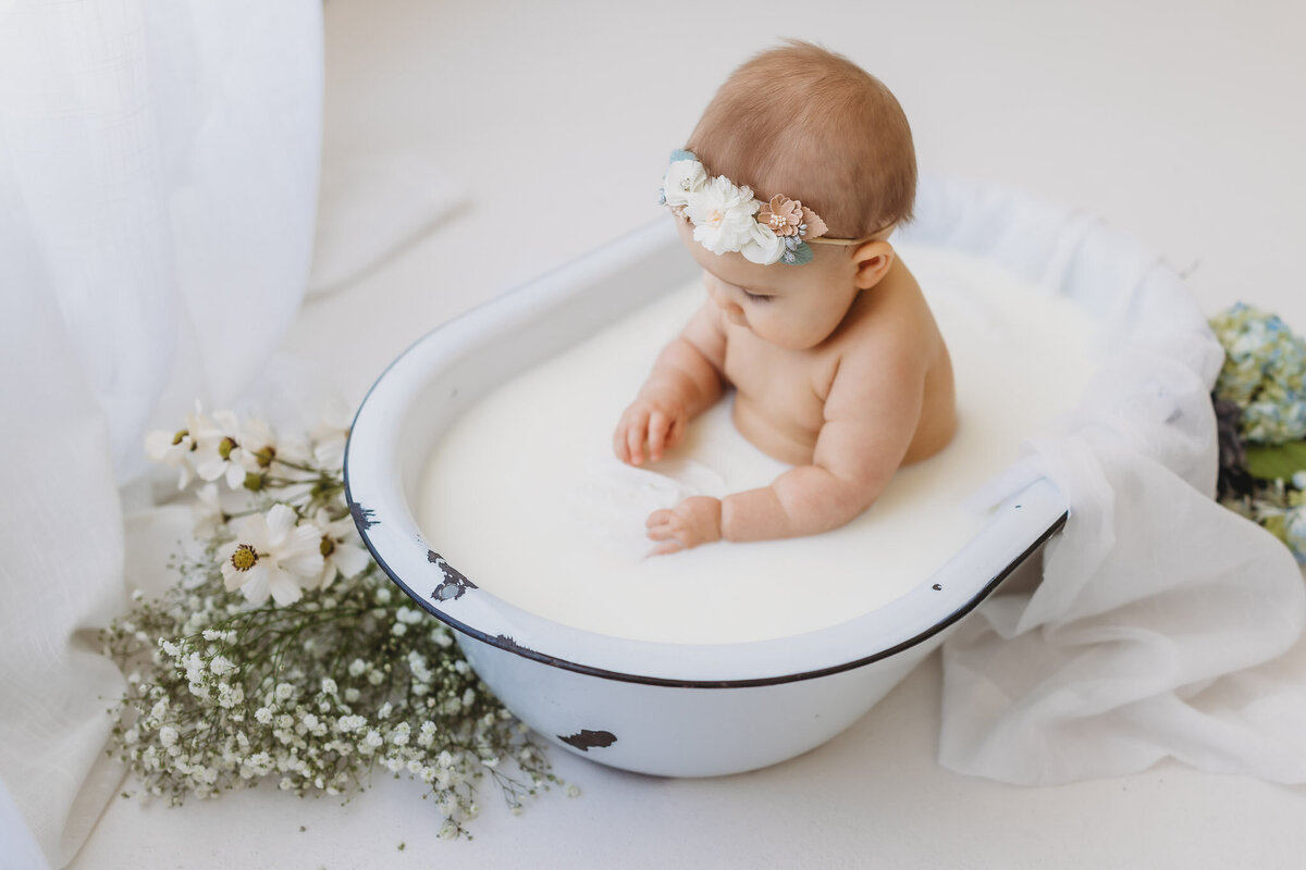 Little baby girl in a small white bathtub with milk splashing surrounded by florals in studio