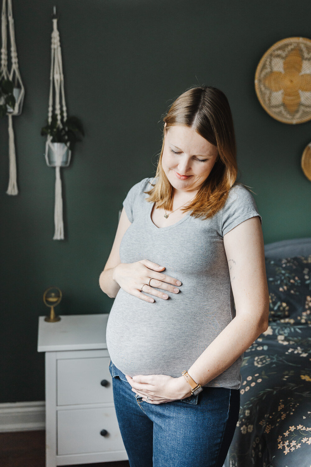 Pregnant women dressed in jeans and grey t-shirt standing in her bedroom. She is lovingly looking down at her pregnant belly.
