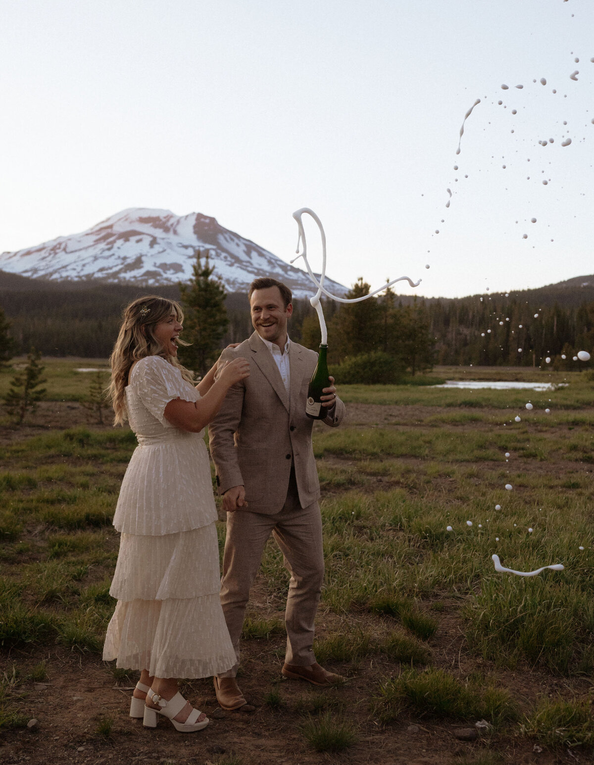 bride and groom stand in an open meadow and pop a champagne bottle as they smile and celebrate their elopement in Bend, Oregon